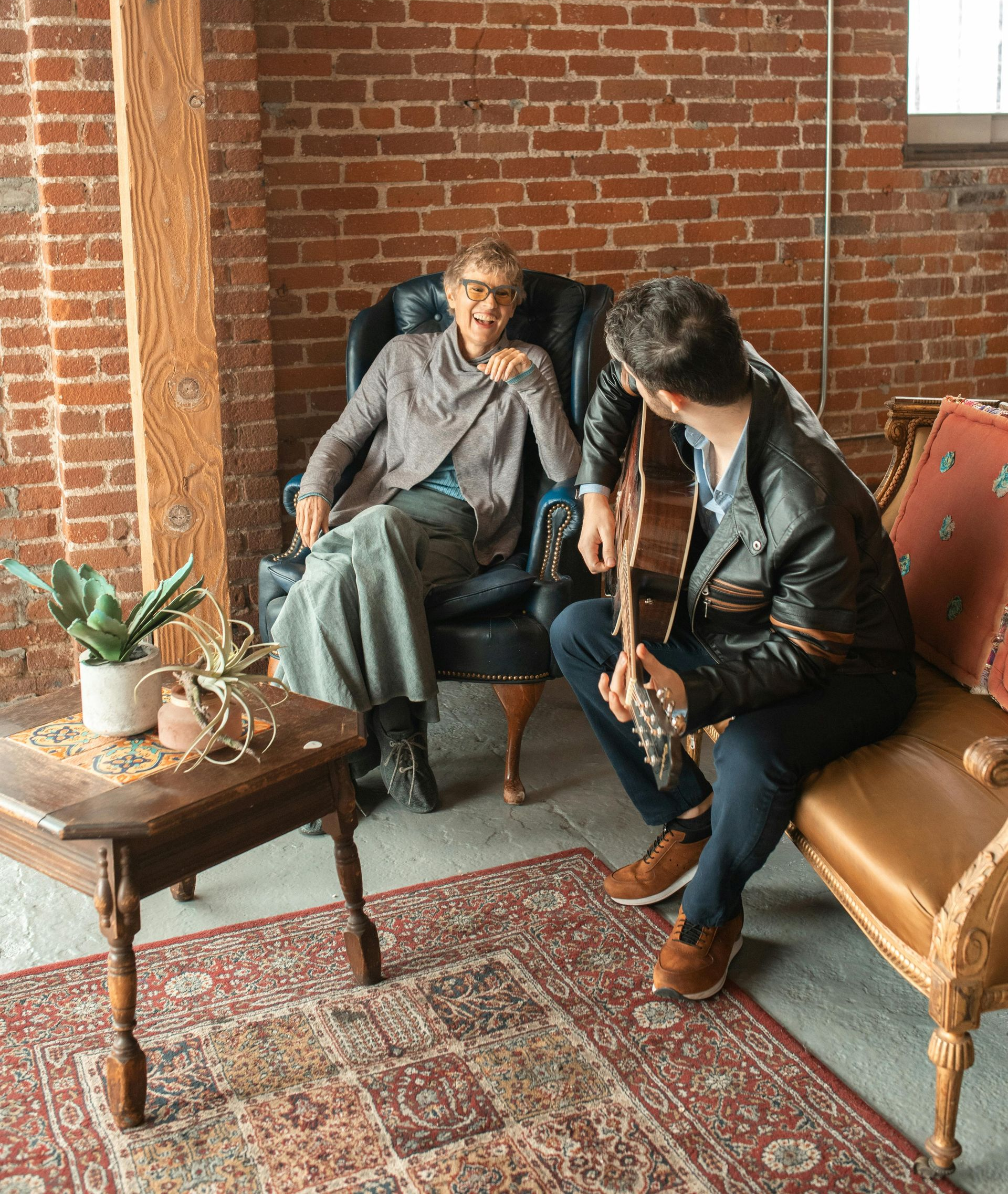 Two people in a brick-walled room. One sits in a chair and smiles. The other sits on a bench and holds a guitar.