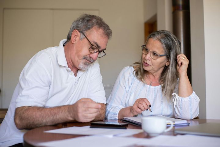 Older couple reviewing documents at a table; man holding small object, woman gesturing, papers and coffee visible.
