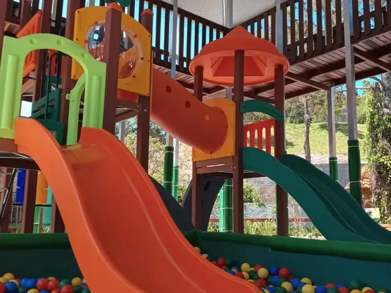 Parque infantil ao ar livre com escorregadores laranja e verde, pequena piscina de bolinhas e estrutura de madeira.