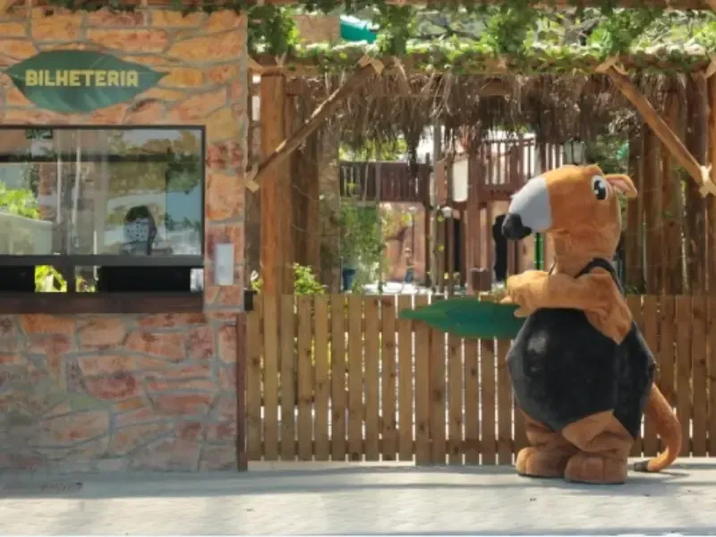 Mascote com traje marrom e preto ao lado de uma bilheteria em um local ao ar livre.