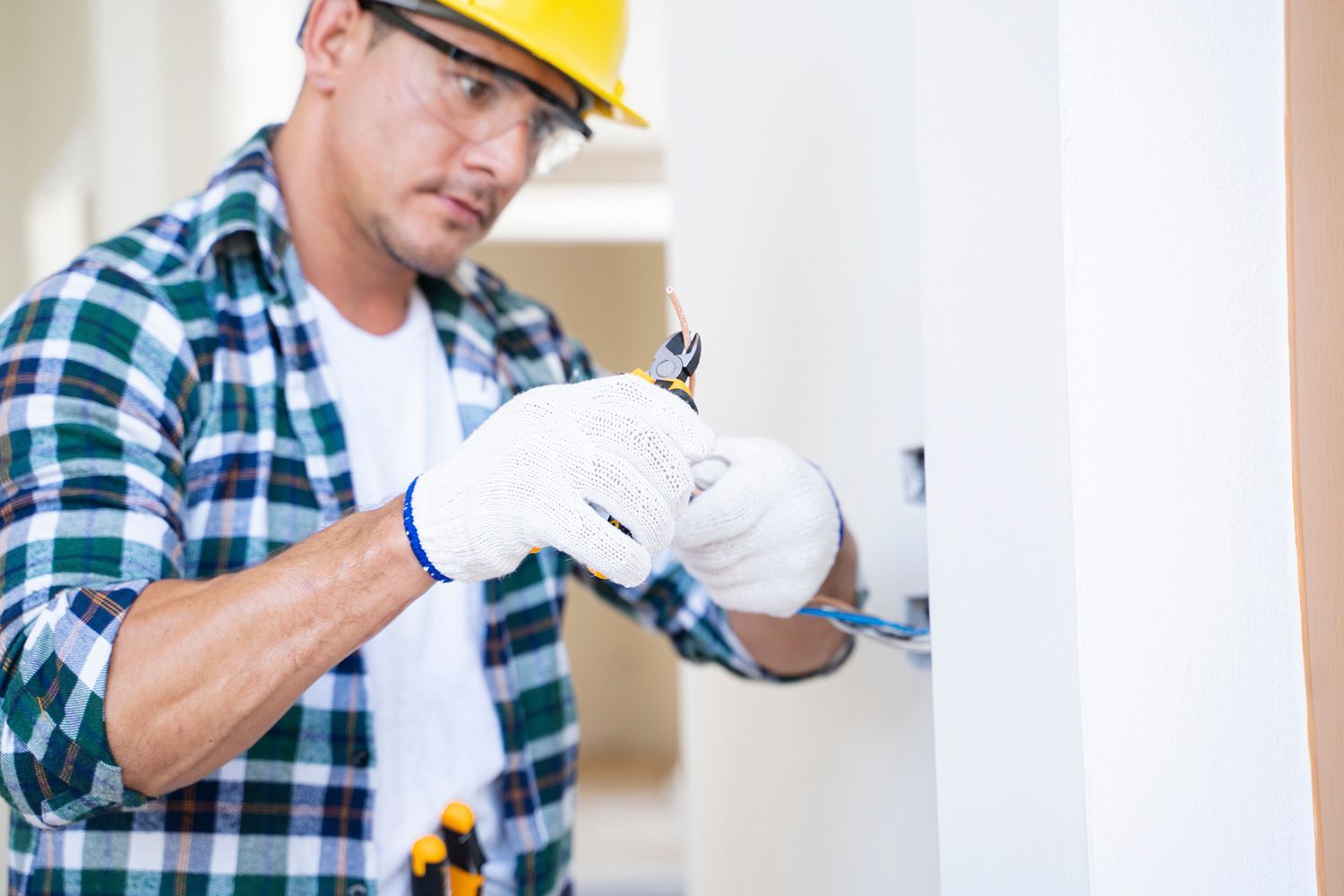 An electrician wearing a yellow hardhat and gloves, with a tool in his hands, rewiring a wall socket