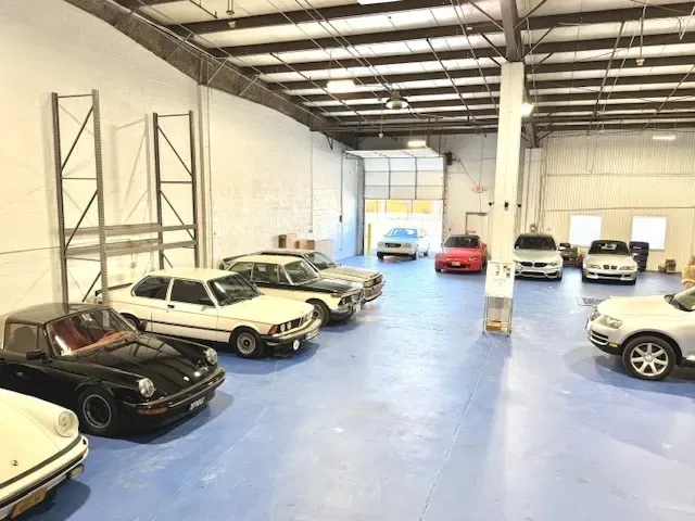 Classic cars parked inside a bright warehouse with a blue floor, open bay door.