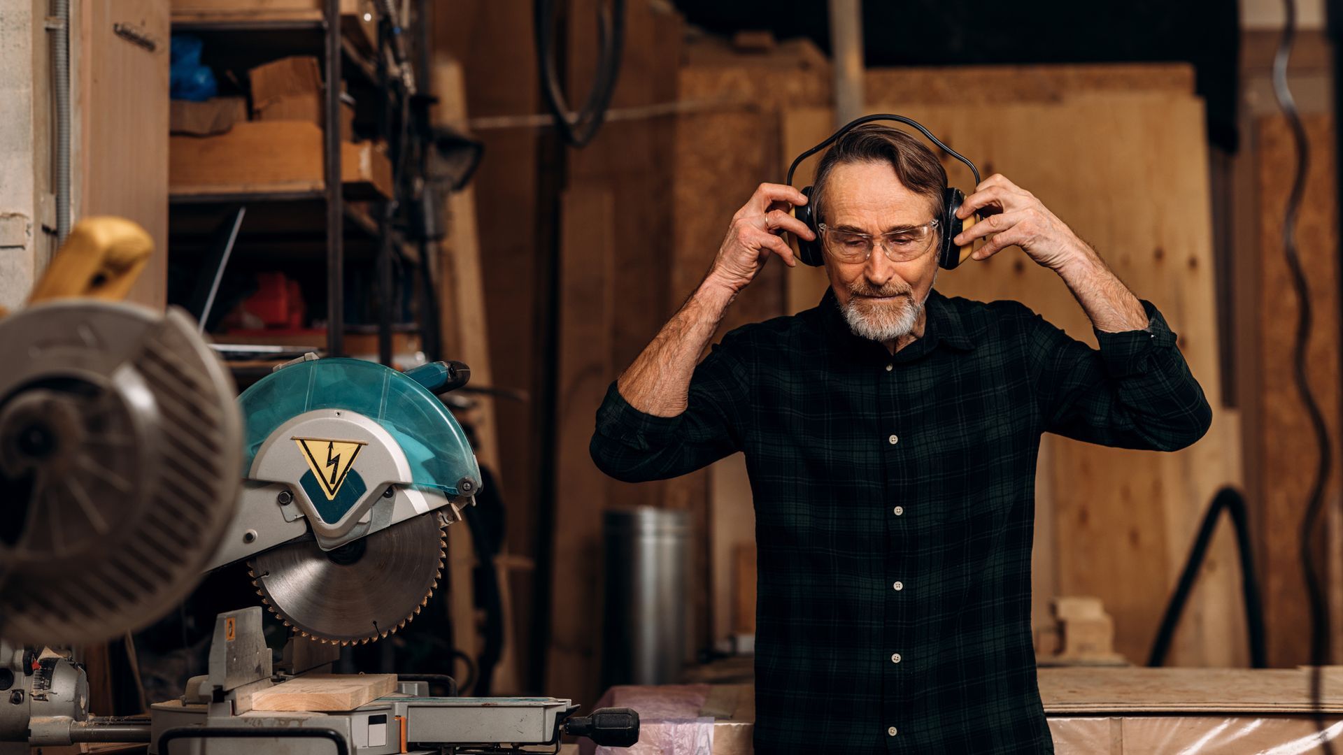 A man wearing headphones is standing in front of a circular saw in a workshop.