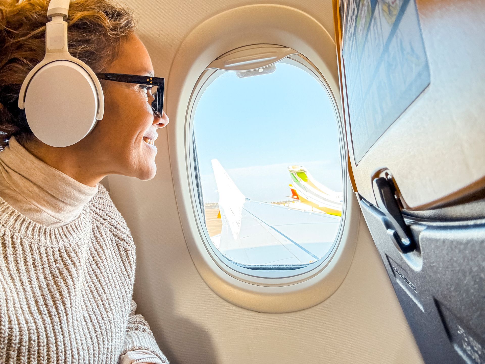 A woman wearing headphones is looking out the window of an airplane.