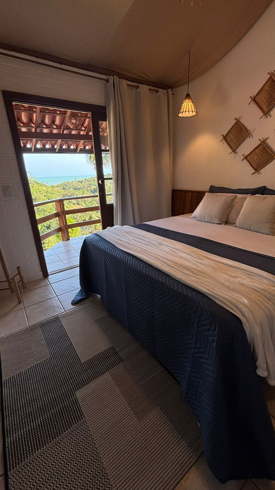Um quarto de hotel com cama, tapete estampado e varanda com vista para uma paisagem verdejante e o oceano ao longe.
