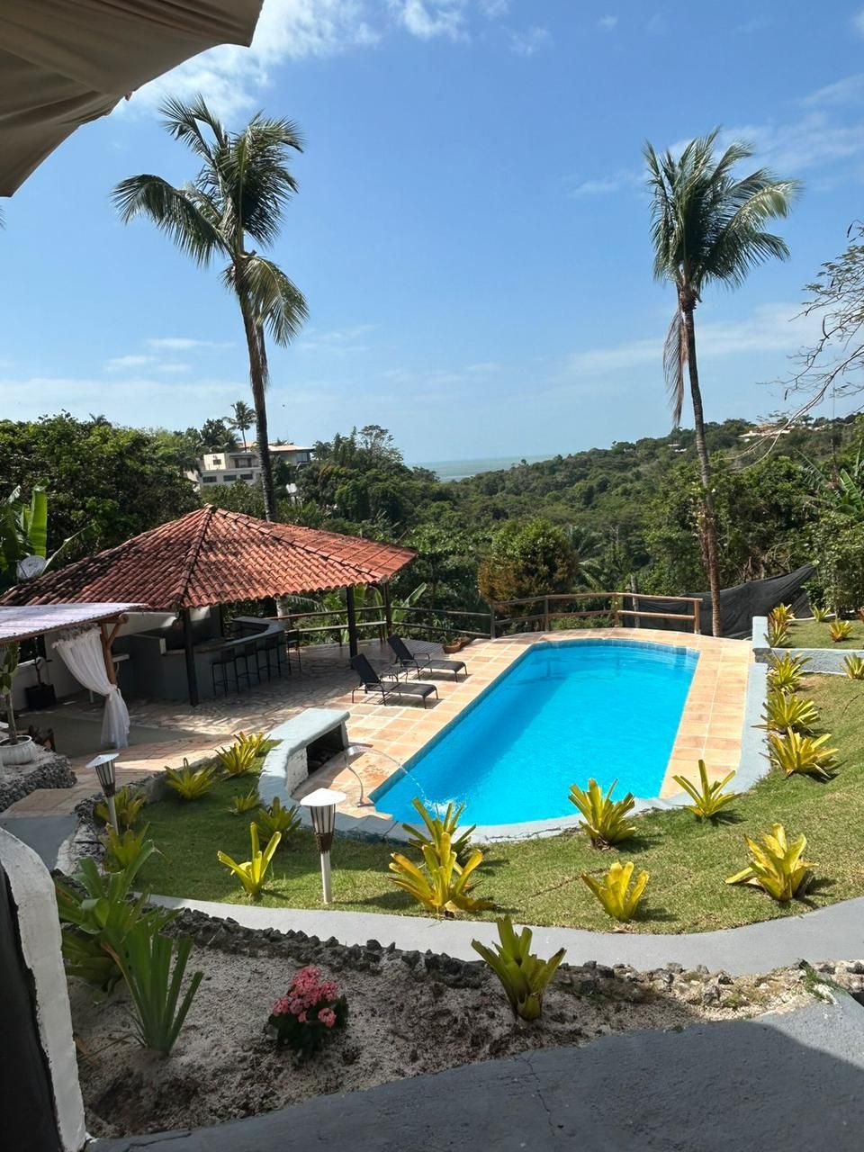 Uma piscina ao lado de um pavilhão com teto de palha, rodeada por árvores exuberantes e com vista para o oceano ao longe.