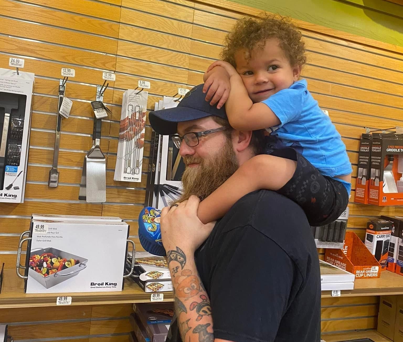 Man with beard and hat carries a child on his shoulders in a store. Child smiles.