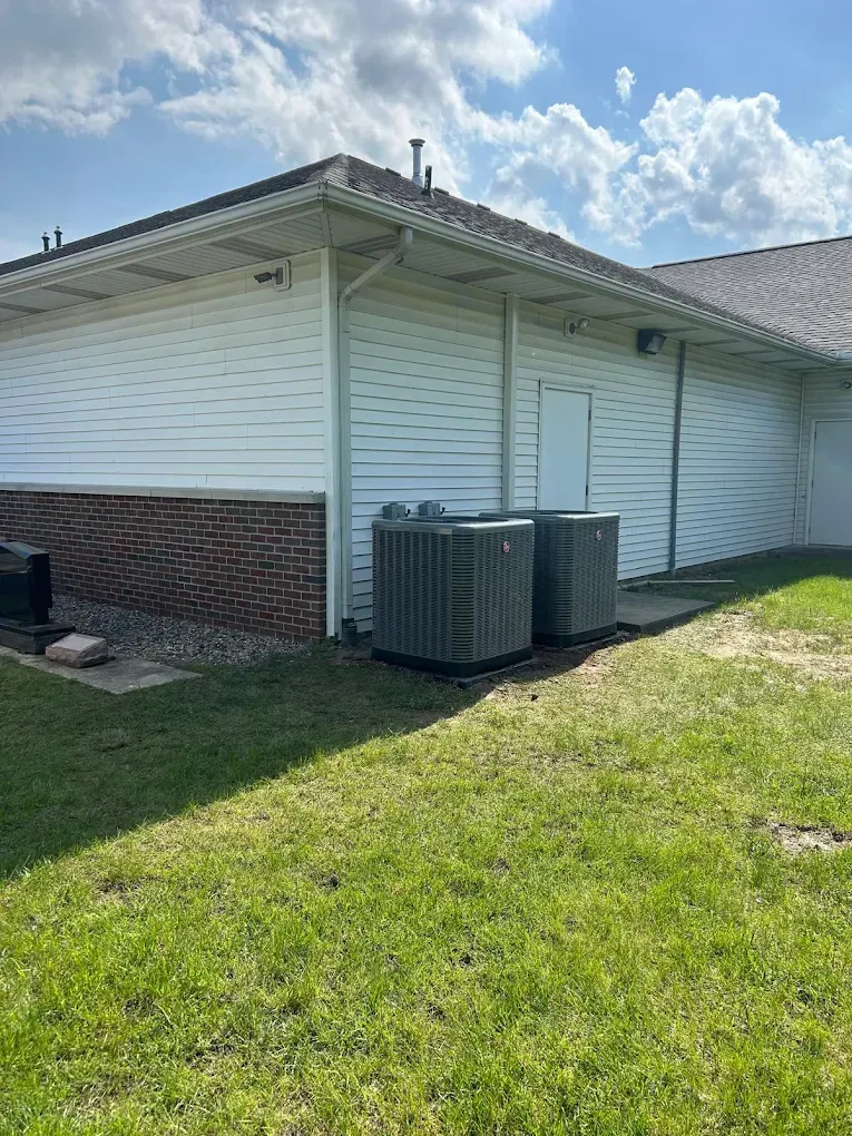 Two air conditioning units sit next to a white building with a brick base on a grassy lawn.