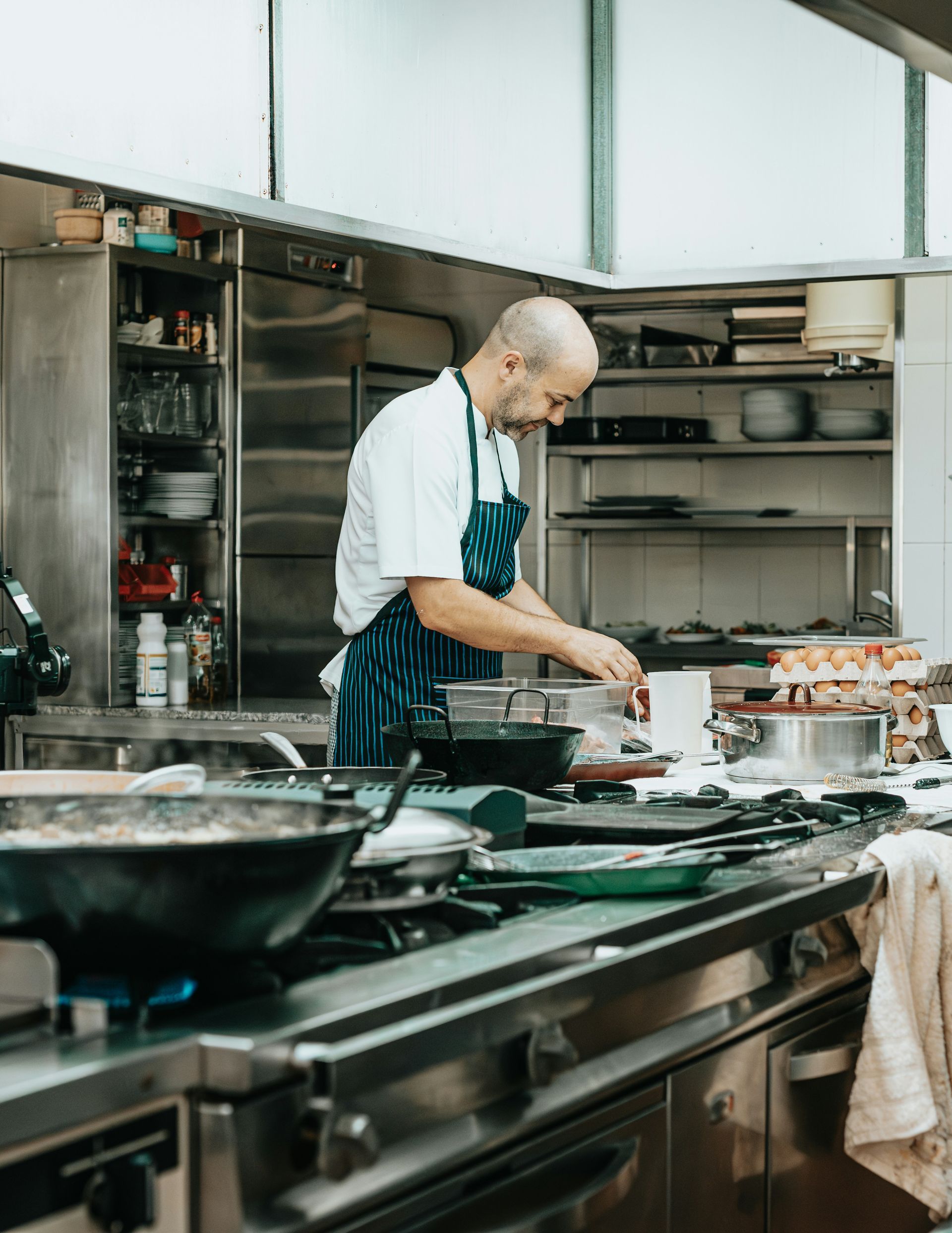 Chef in a commercial kitchen, wearing apron, cooking on a stovetop with various pots and pans.