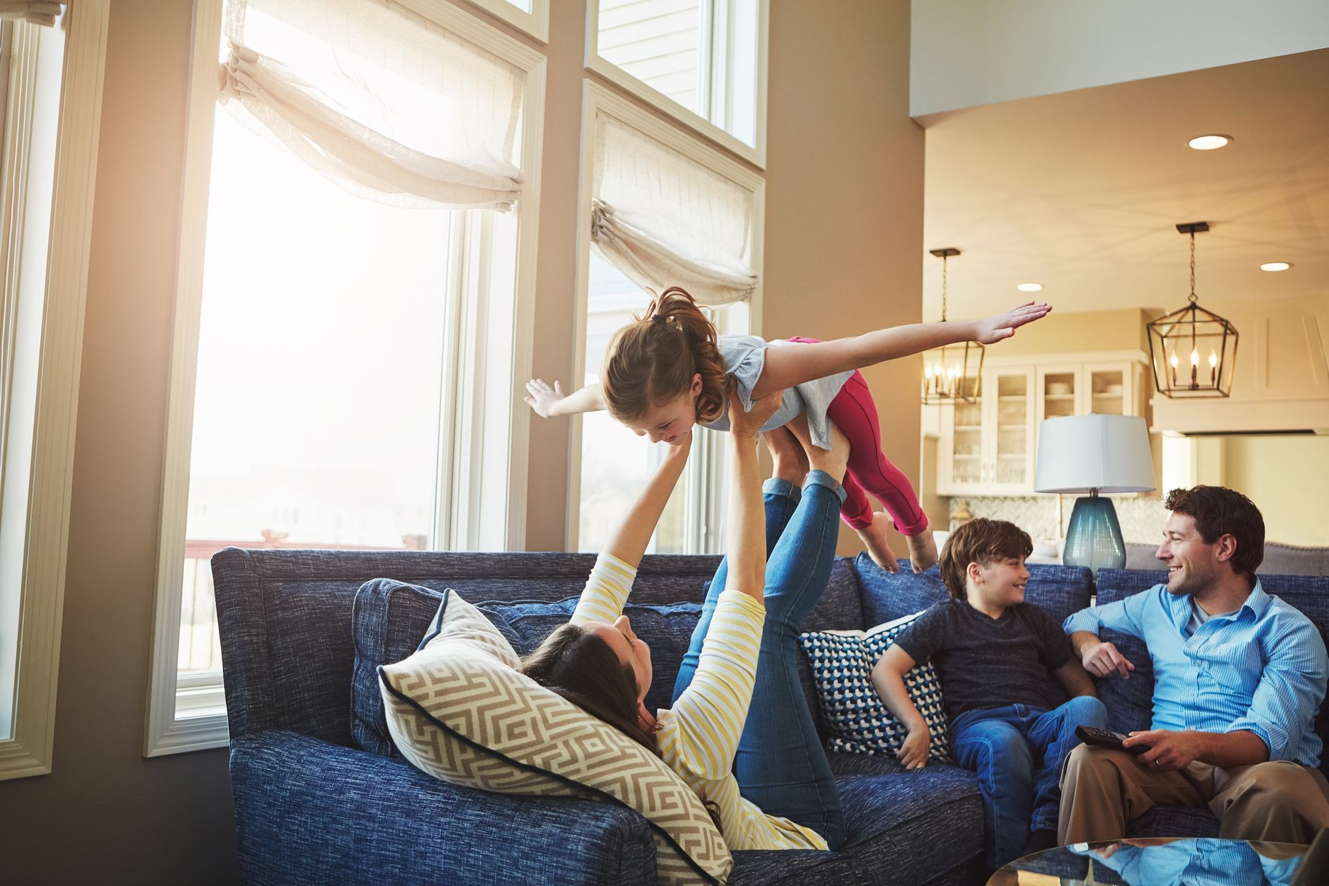 Family playing in a living room. A woman holds a child in the air, a boy watches, and a man sits.