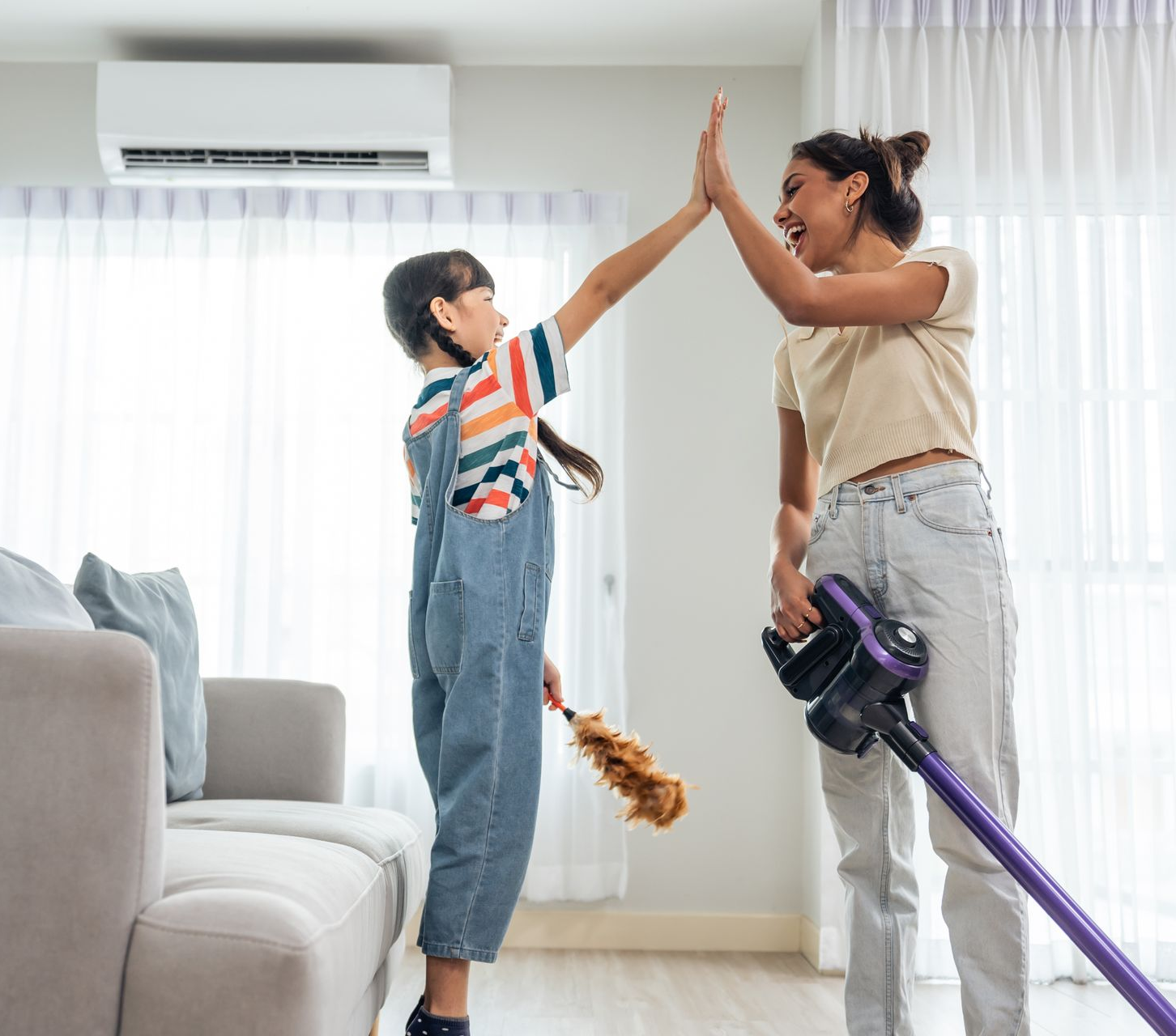 Woman and child high-five after cleaning. Child holds duster, woman has vacuum in living room.
