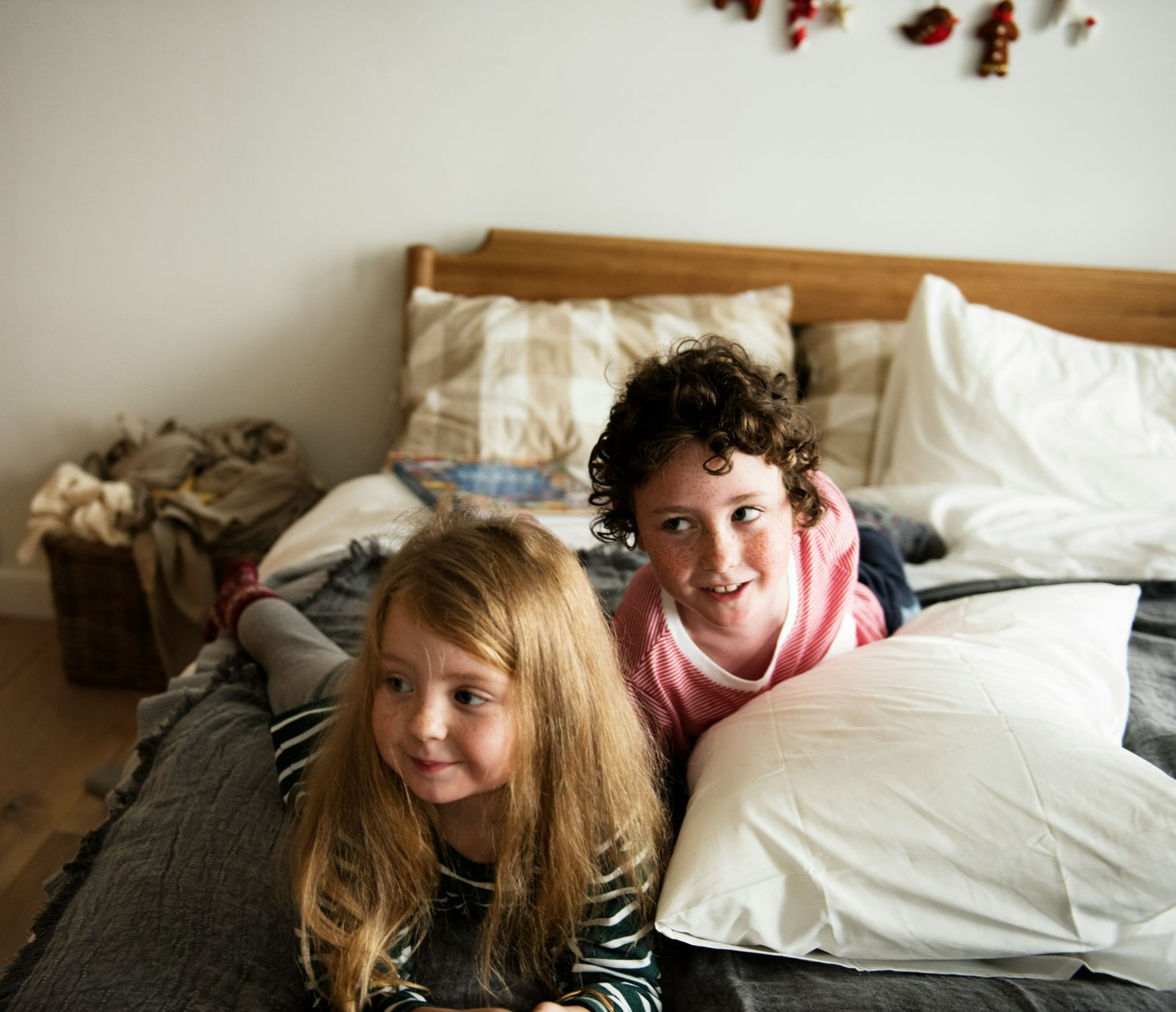 Two children lying on a bed, smiling. One has long blonde hair, the other has curly hair.
