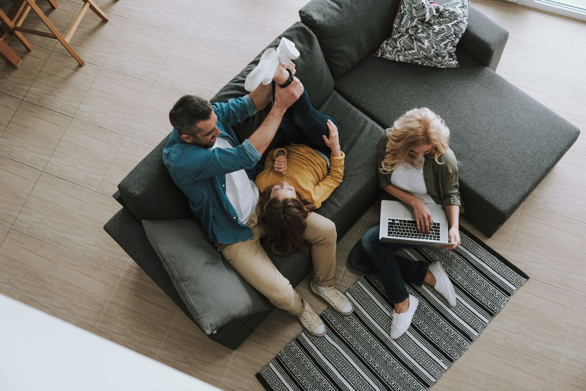 Family on a couch: man playing with a child, woman using laptop. Neutral tones, living room setting.