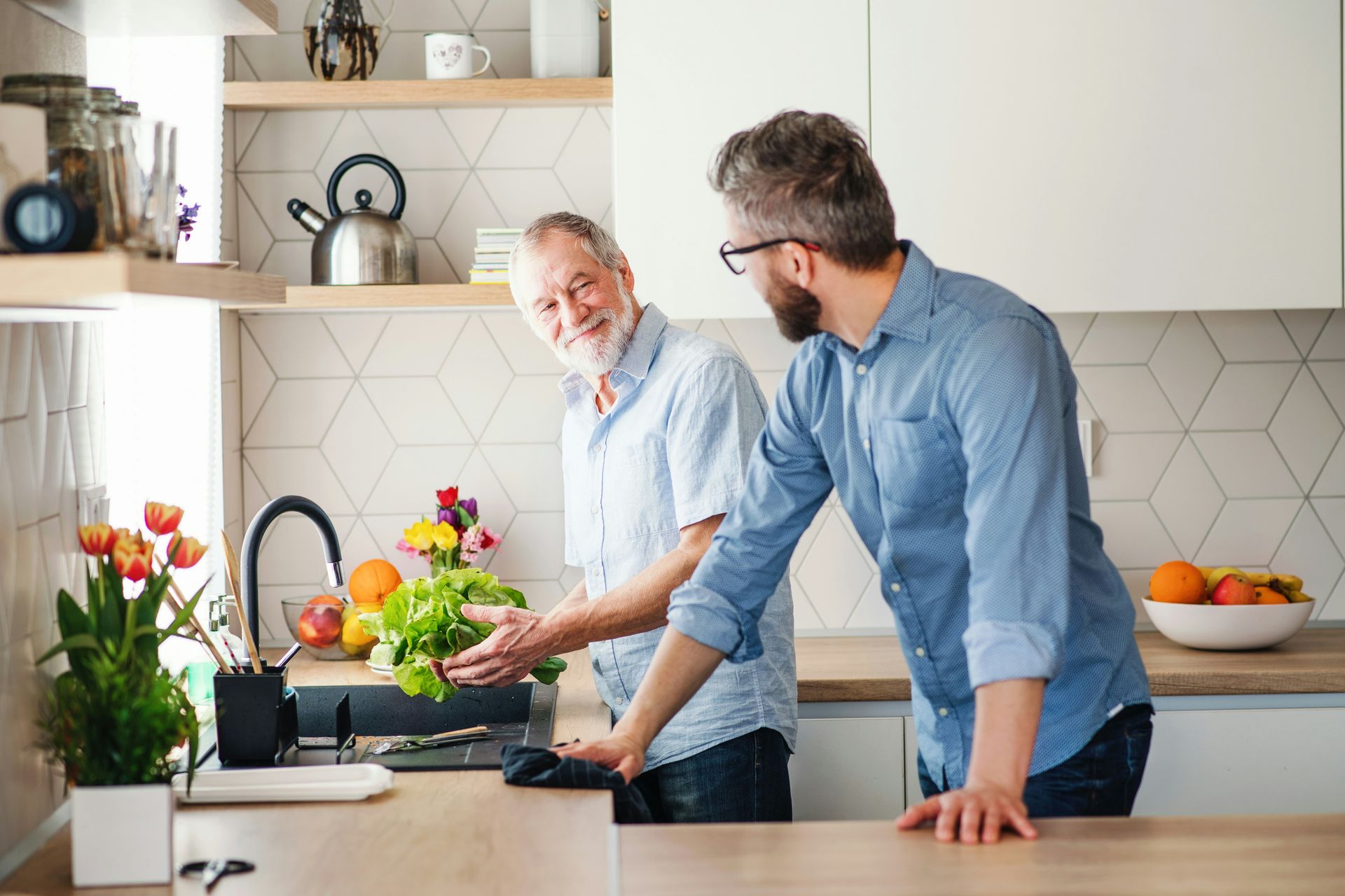 Two men in a kitchen, washing lettuce. One smiles while the other looks on.