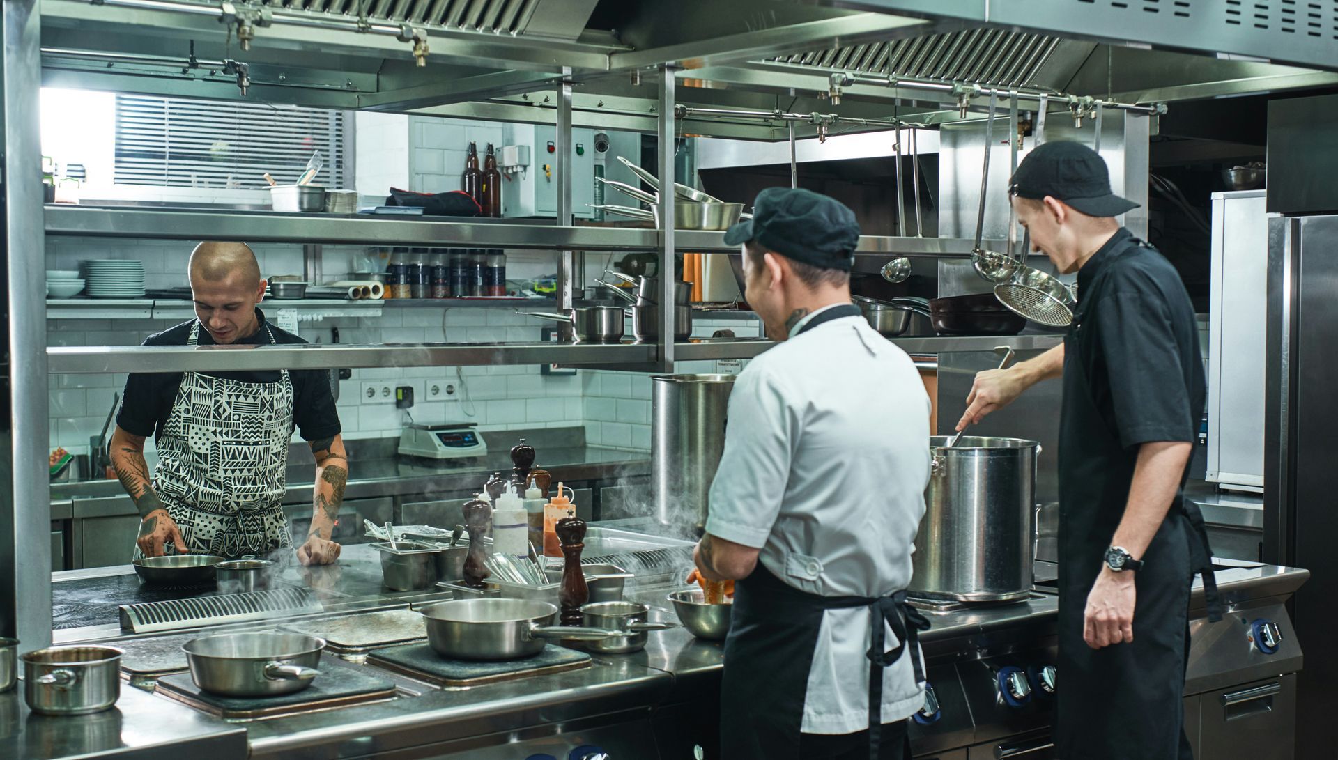 Chefs working in a commercial kitchen; one stirring a pot, another looking at a pan, another at a station.
