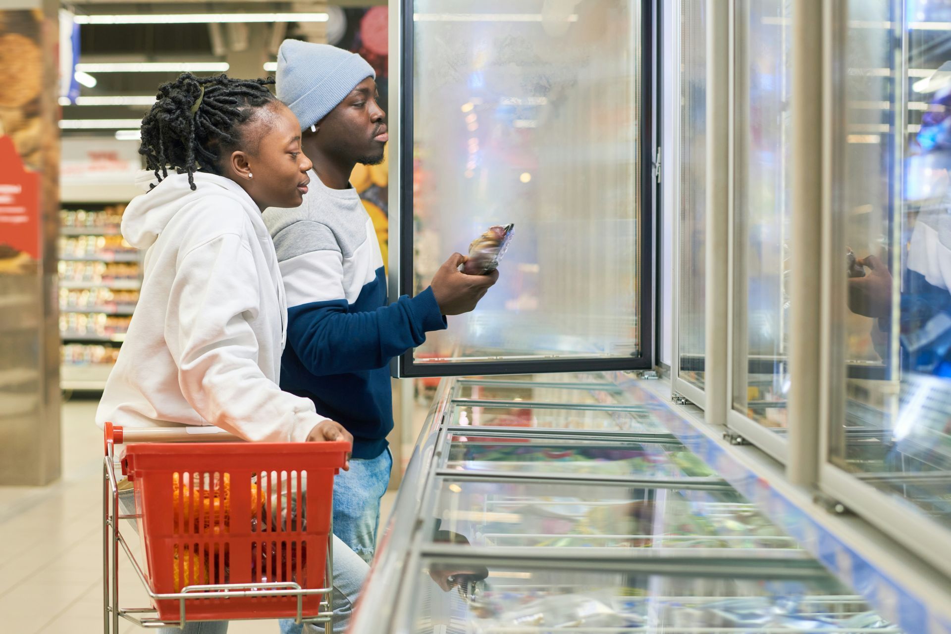 Two people at a grocery store, looking at frozen food in a freezer. One holds a package. Red shopping basket.