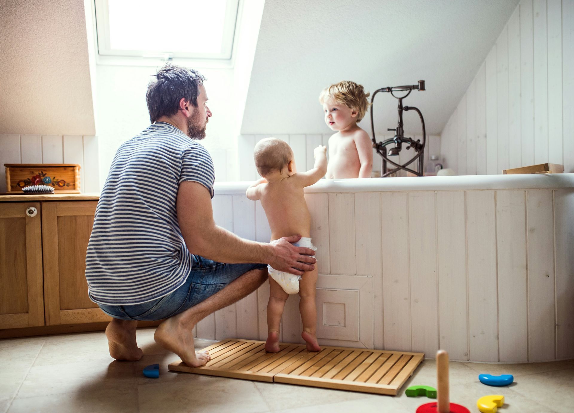 Man helping two toddlers in a bathtub. One child is in diapers and the other is standing. Brightly lit room.