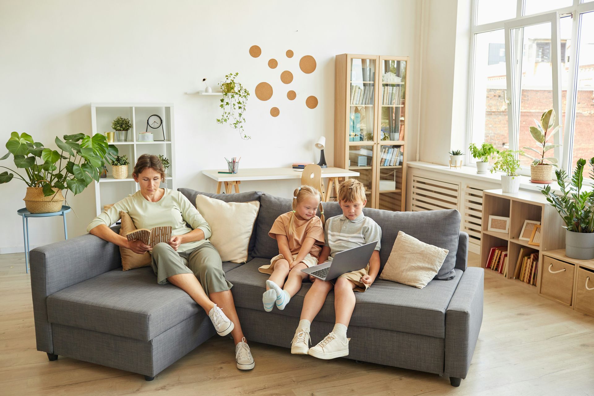 Family on a gray sofa in a light-filled living room; person using a tablet, kids using a laptop, plants.