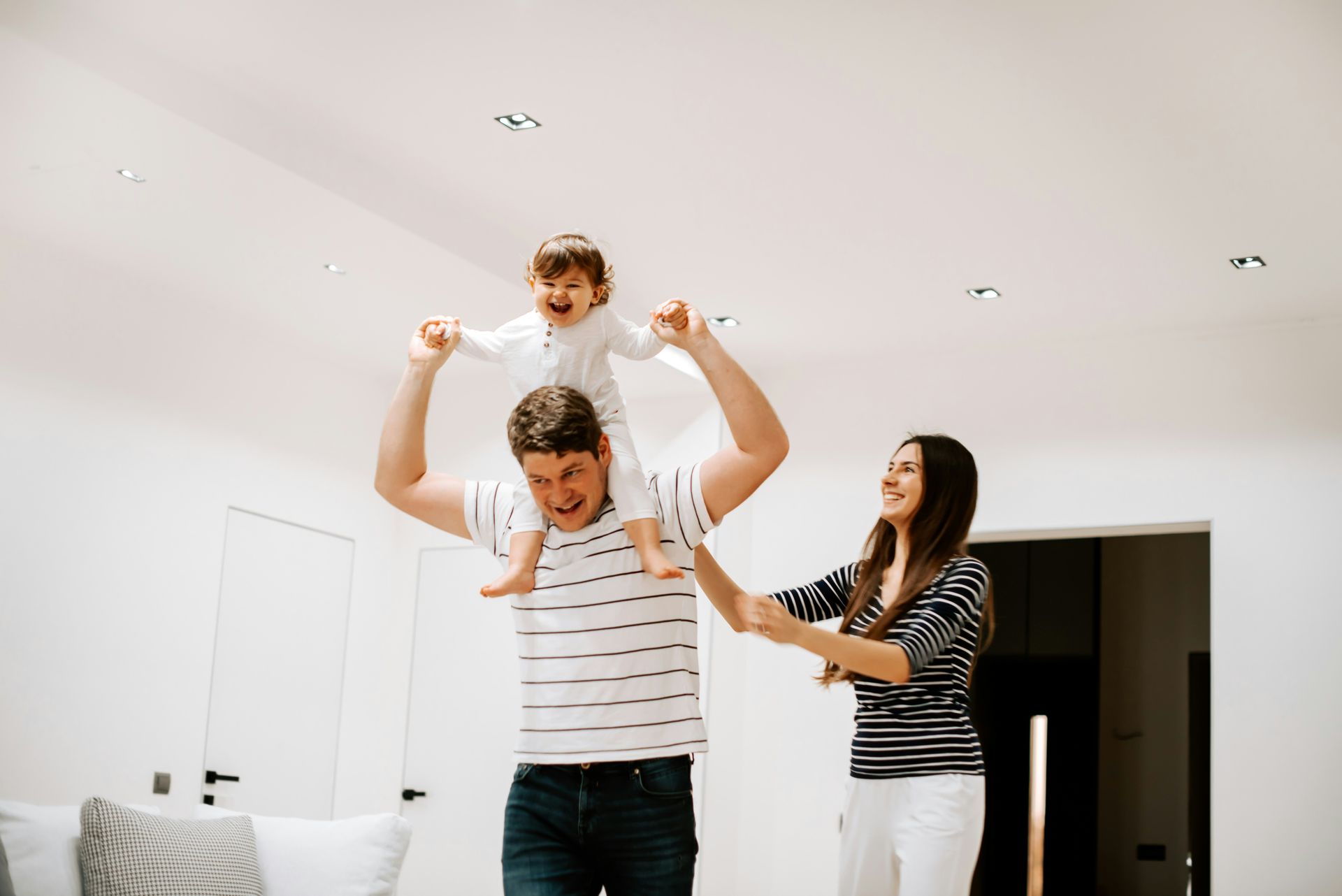 Father carrying child on shoulders, smiling, with mother beside them. Brightly lit, modern interior.