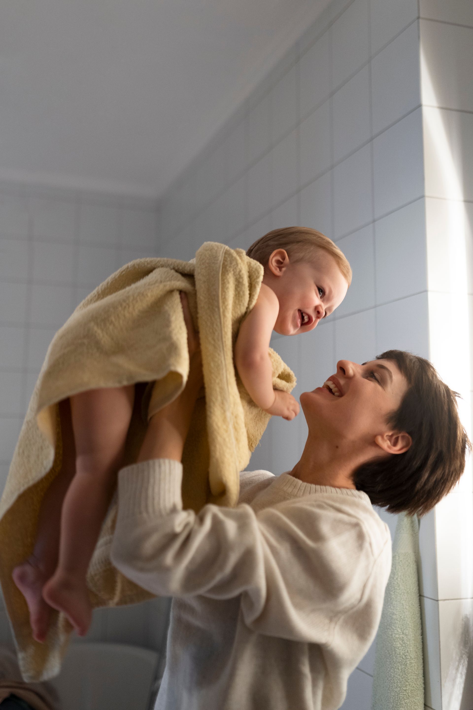 Woman holding a laughing baby wrapped in a towel in a bathroom with tiled walls.