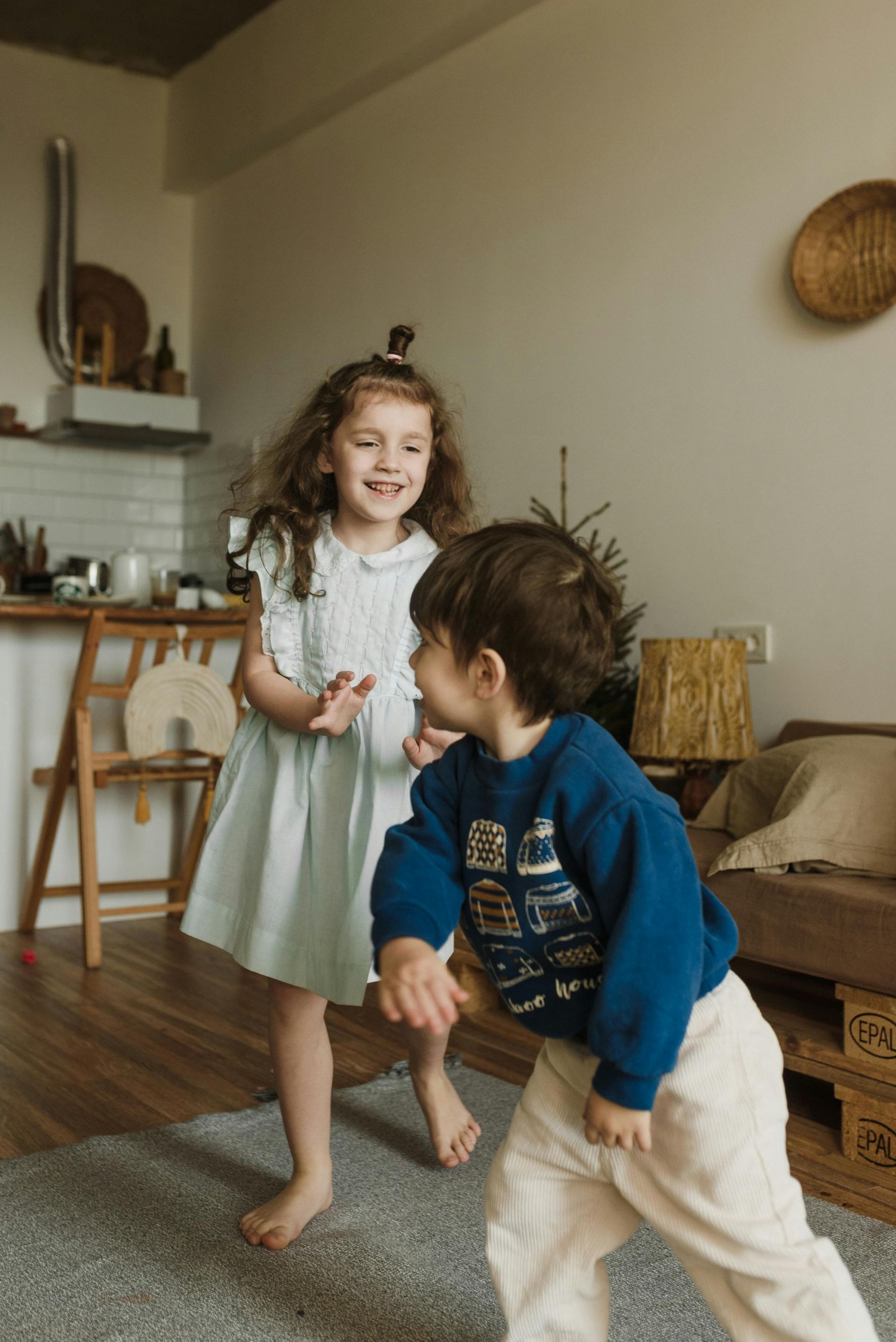 Two children smiling, dancing in a room. Girl in blue dress, boy in blue sweater and tan pants.