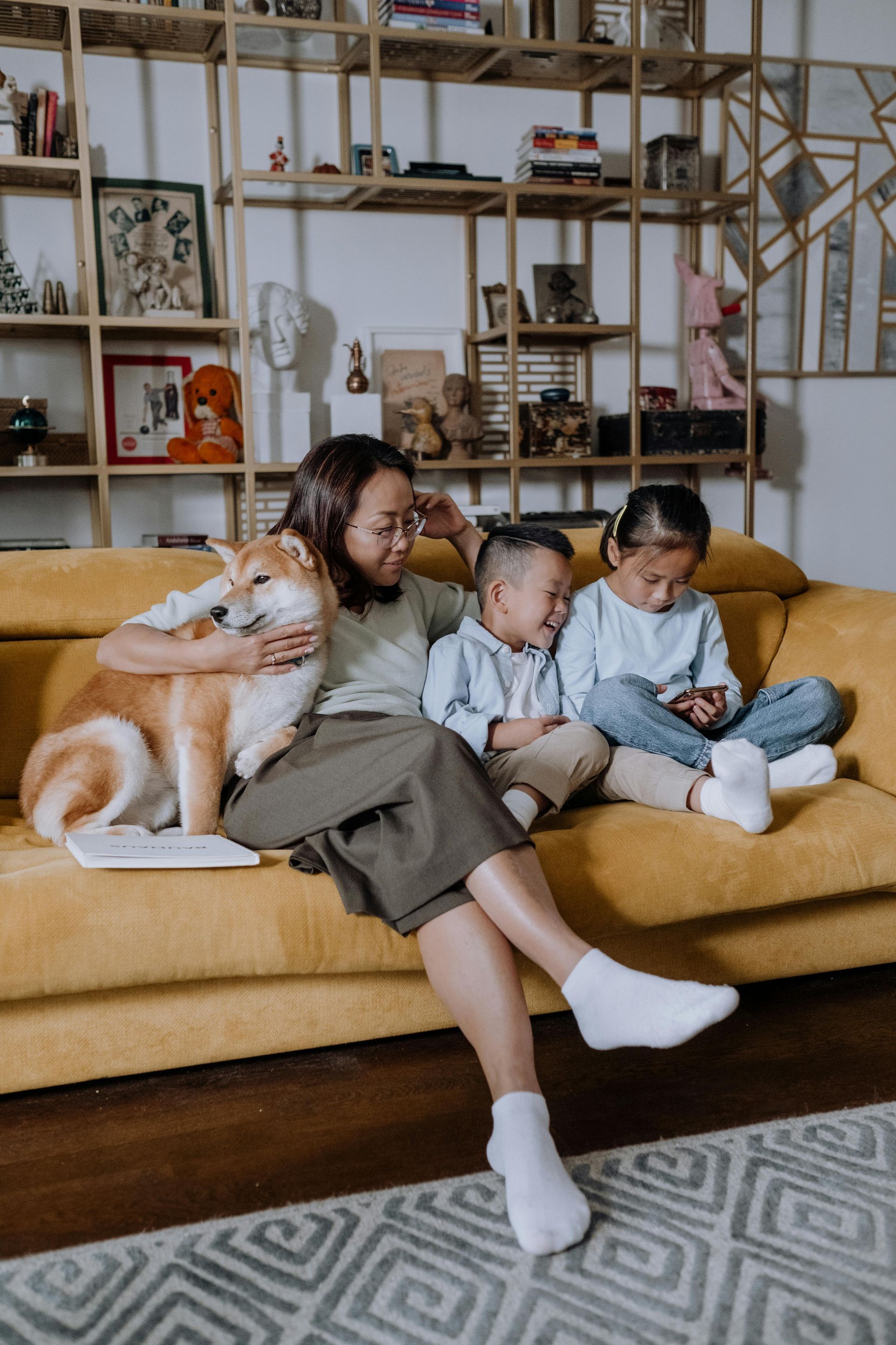 Woman, two children, and Shiba Inu dog on a yellow sofa in a living room, smiling.
