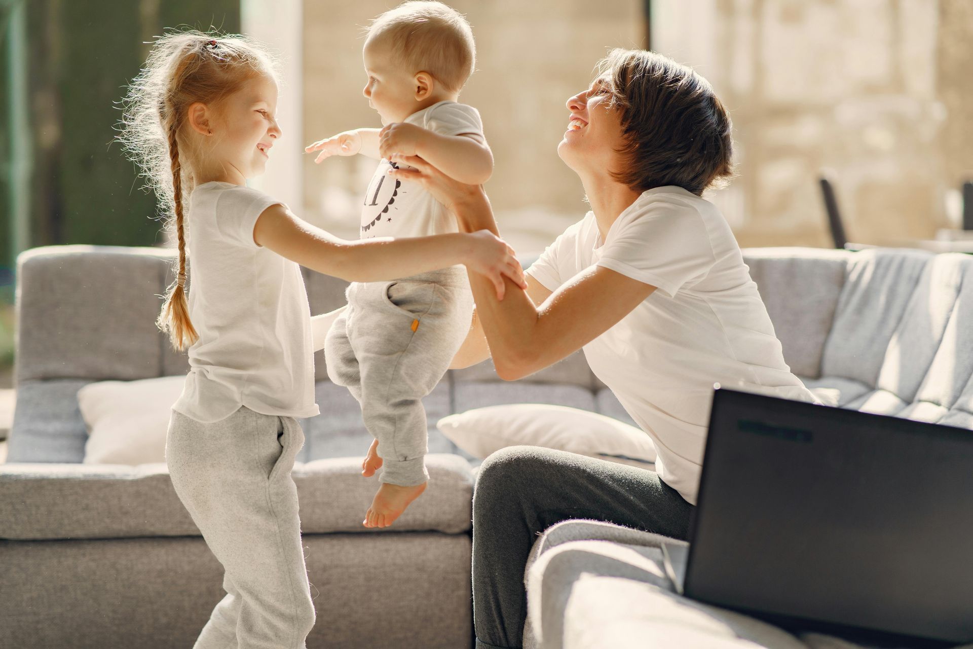 Woman lifting baby, another child smiles, seated near a laptop on a couch in a bright room.