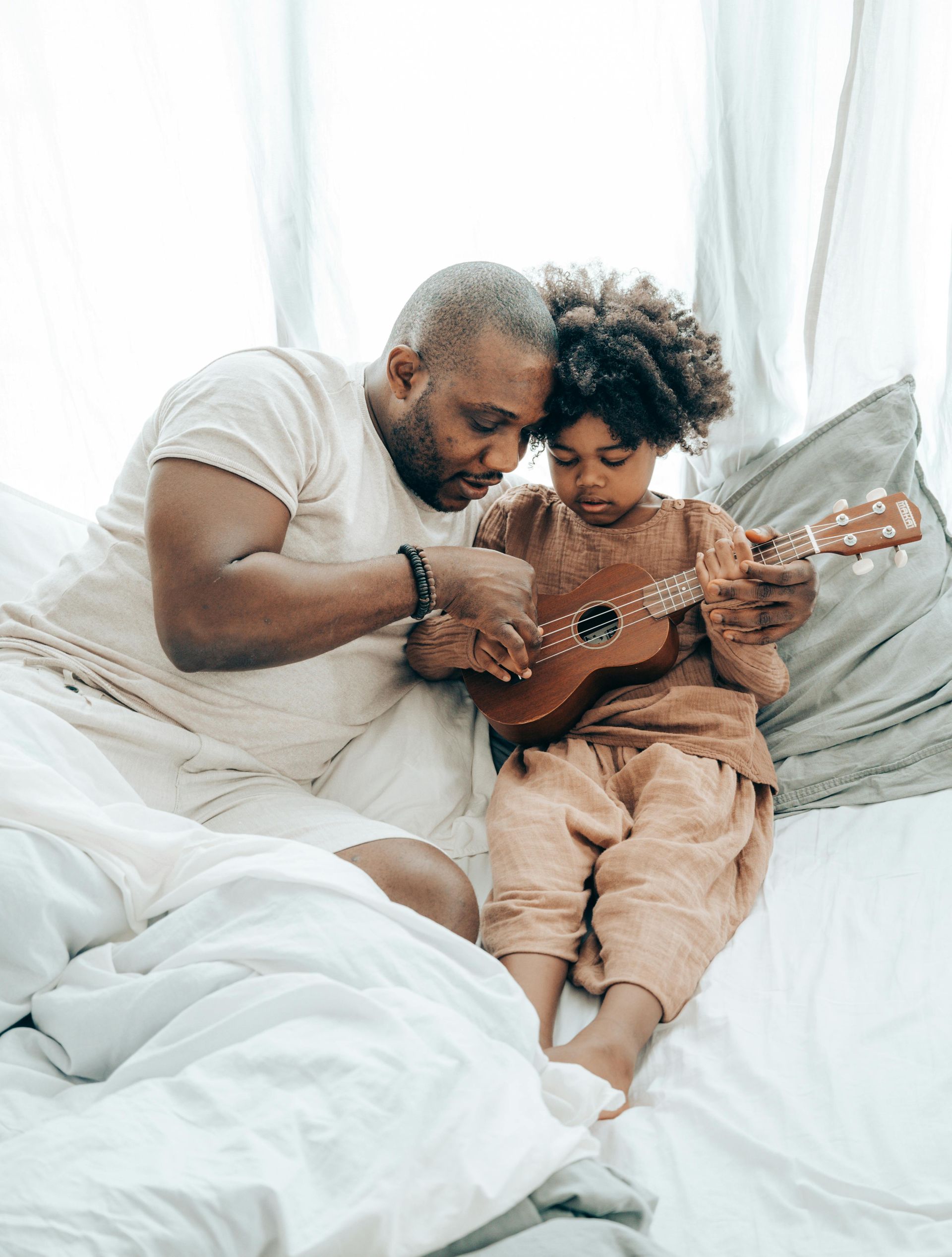 Father and child playing ukulele together on a bed.