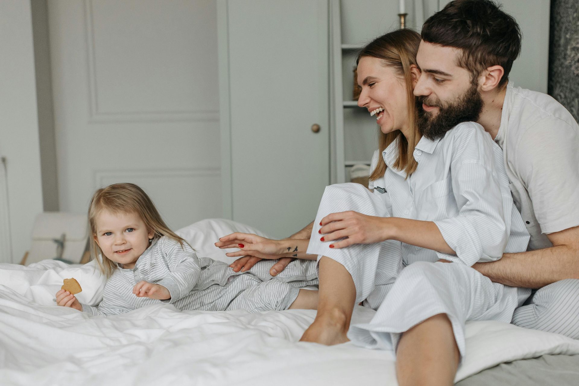 Family on a bed, smiling. Child holding a snack. Parents in pajamas, laughing. Bedroom setting.
