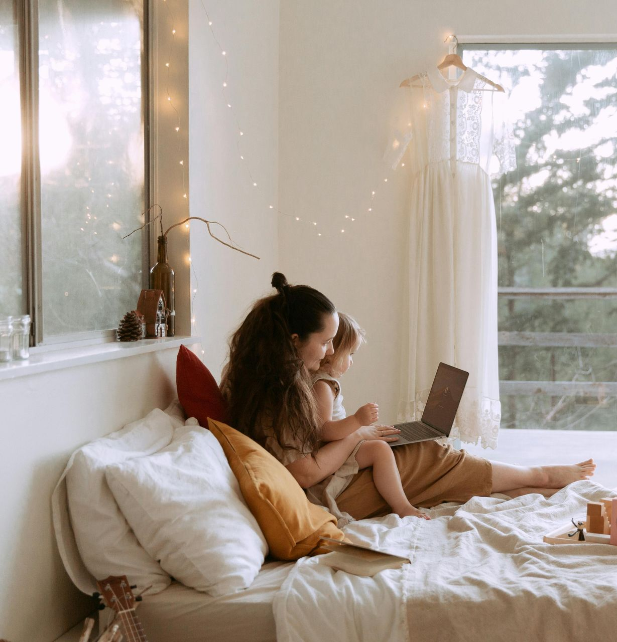 Woman and child on a bed looking at a laptop. The room is bright with a window and a dress hanging up.