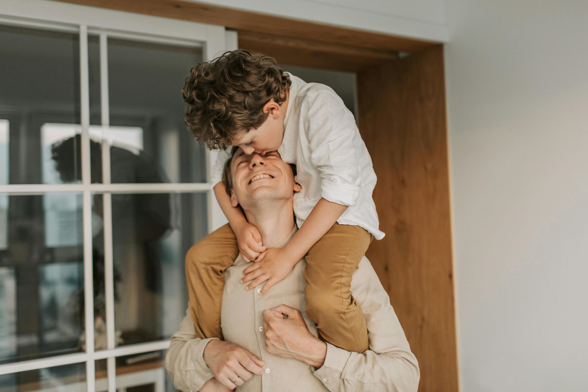 Boy kisses father's head while sitting on his shoulders near a window.