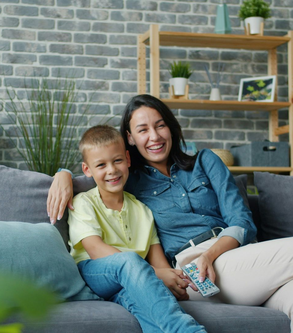 Woman and child on couch, smiling. Living room with plants and shelving.