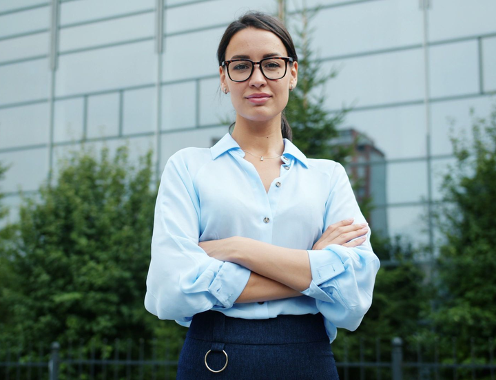 Person in a light blue shirt stands with arms crossed in front of a glass office building.