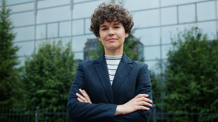Person with curly hair in navy blazer stands with arms crossed in front of a glass building.