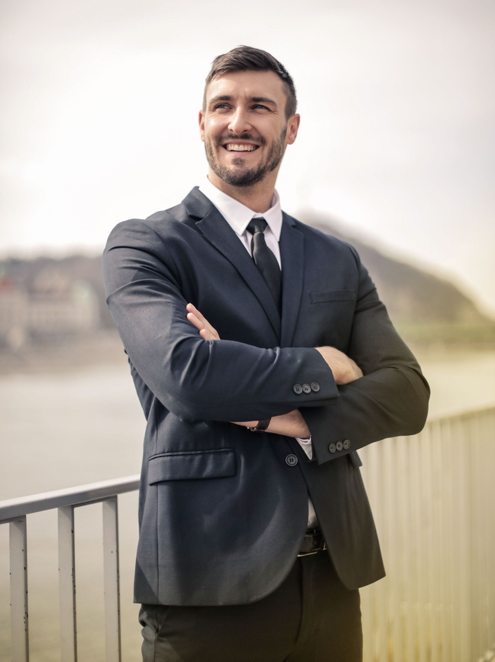 Smiling man in a dark suit stands with arms crossed on a balcony outdoors.
