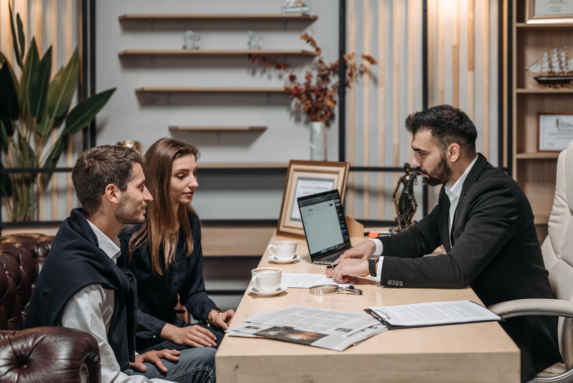Two people meeting with a suit-clad advisor at a desk, reviewing documents and a laptop in a modern office.