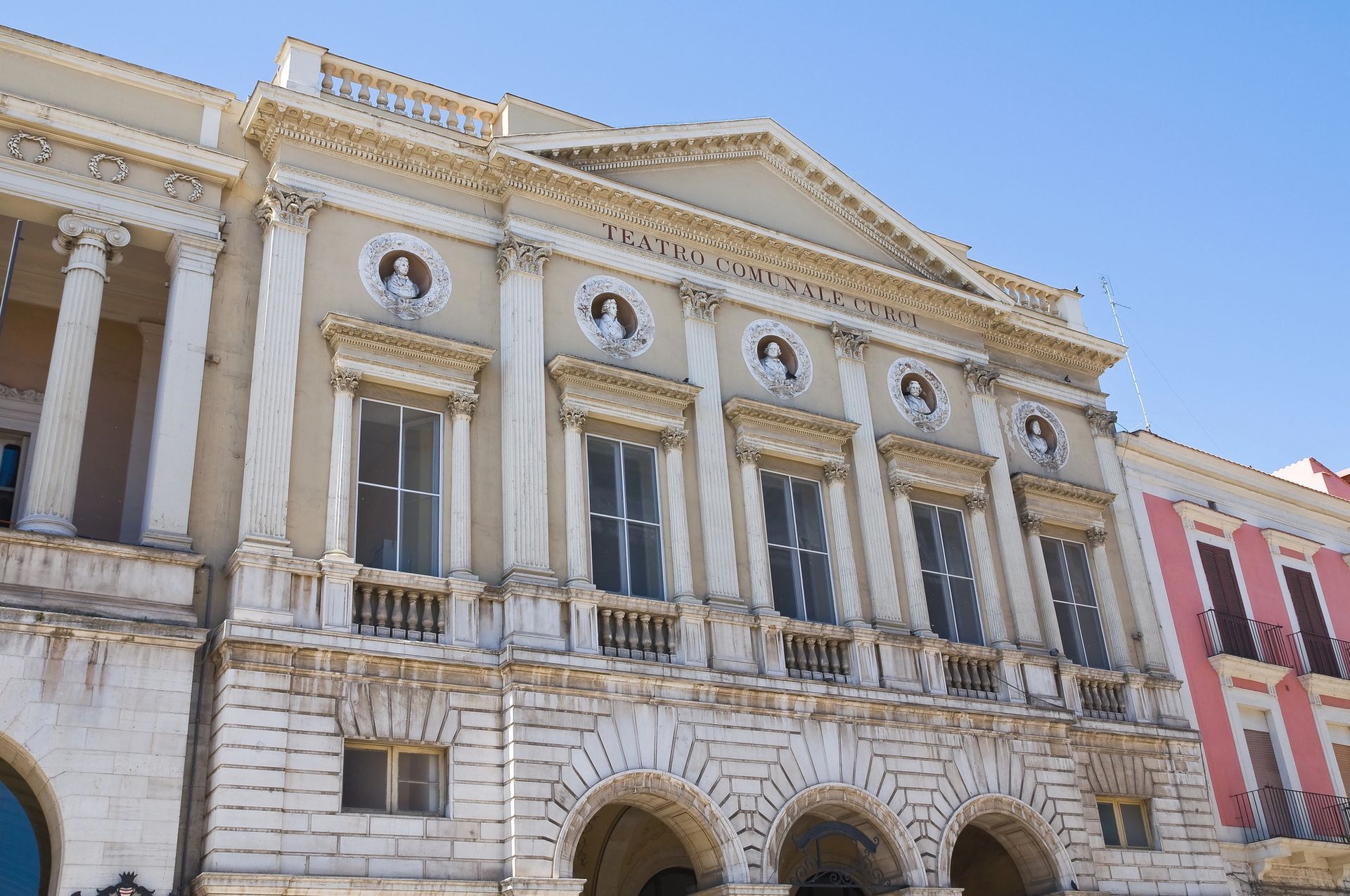 Elegante edificio neoclassico bianco con colonne e ingressi ad arco, affacciato su una via cittadina.
