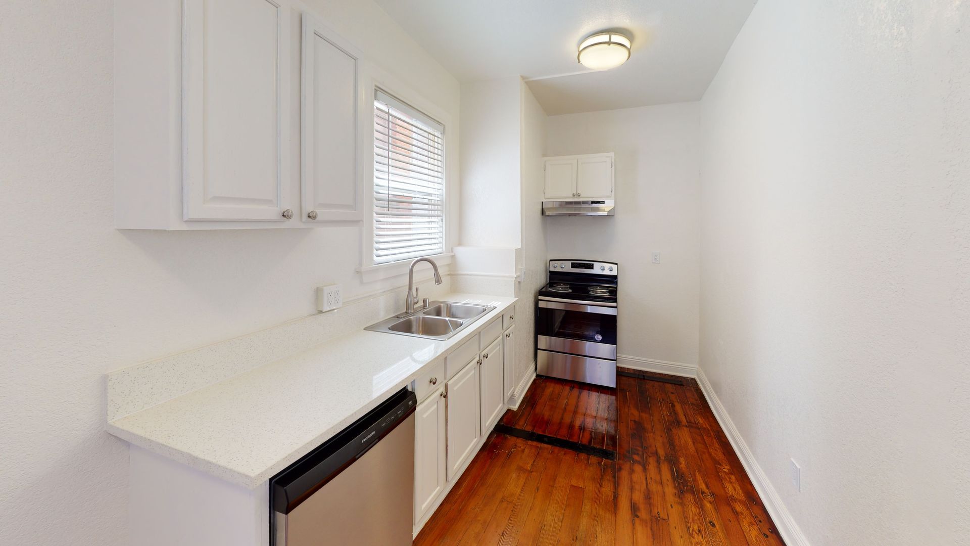 Small, bright kitchen with white cabinets, countertops, and appliances. Wooden floor and a window.