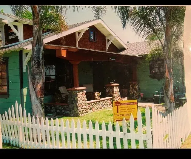 Green craftsman house with white picket fence, stone pillars, and palm trees. A sign is in the yard.