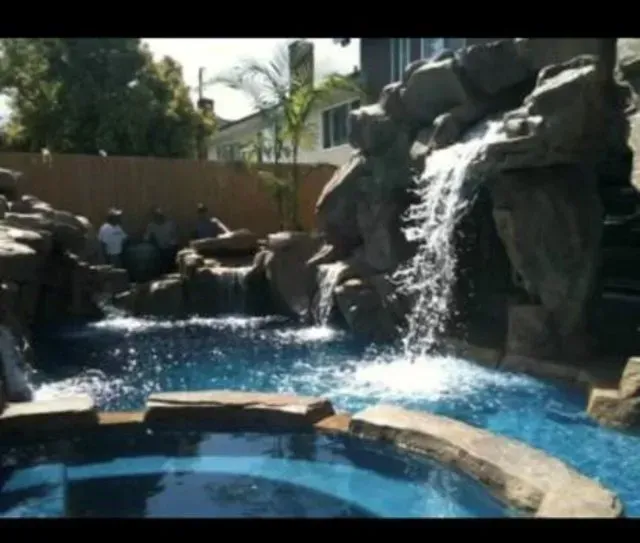 Waterfall cascading into a backyard pool. Rocks surround the pool; a few people stand nearby.