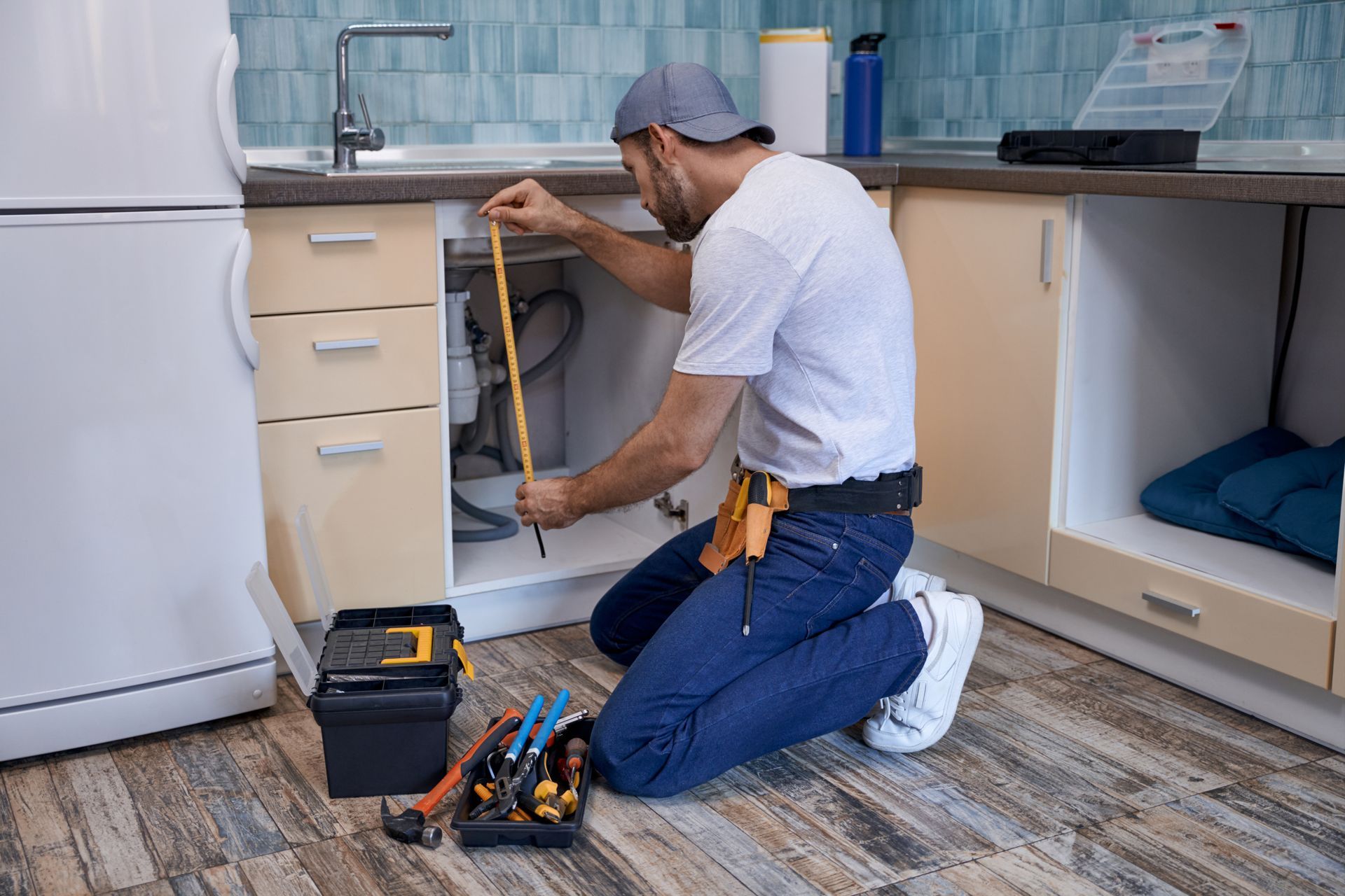 Plumber measuring under a kitchen sink. He is kneeling, wearing a tool belt and hat, with tools nearby. Plumber measuring under a kitchen sink. He is kneeling, wearing a tool belt and hat, with tools nearby.