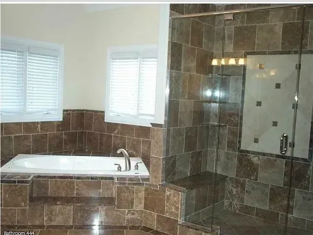 A bathroom with a tiled shower and tub. Large brown tiles are on the walls. Two windows are present.