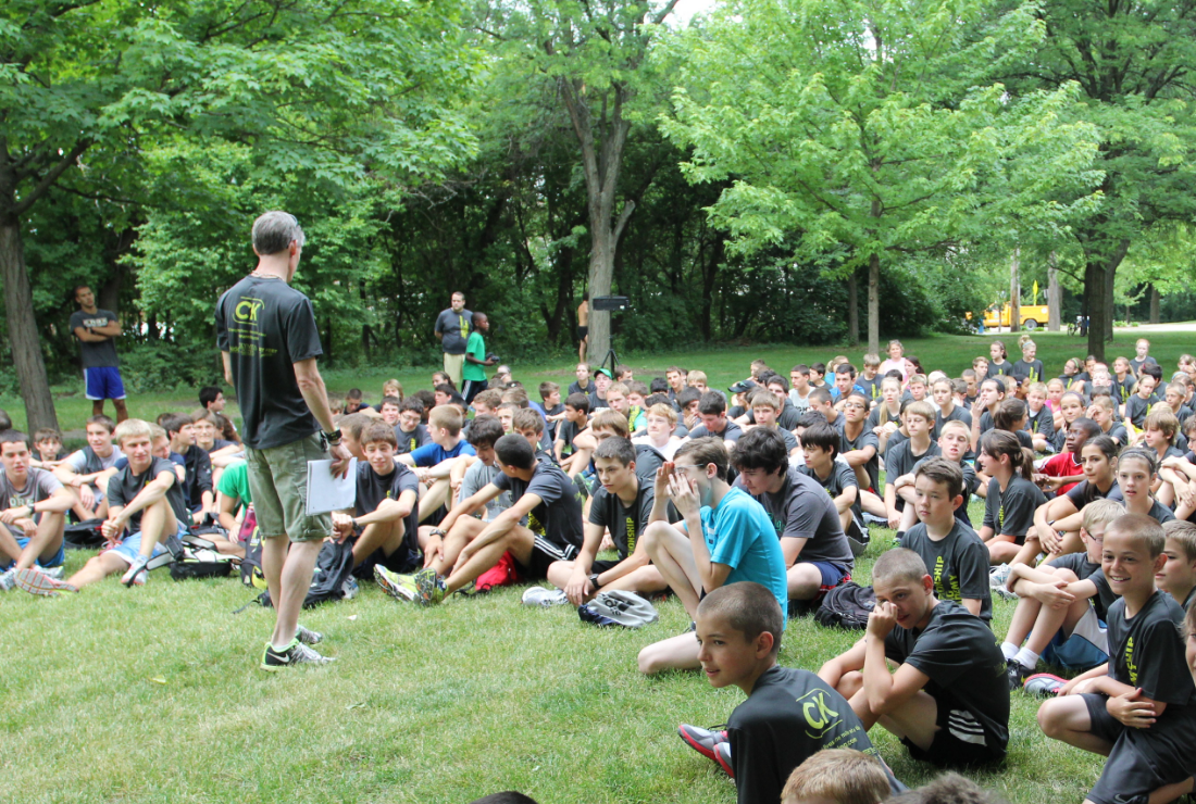 A man is standing in front of a large group of people sitting on the grass.