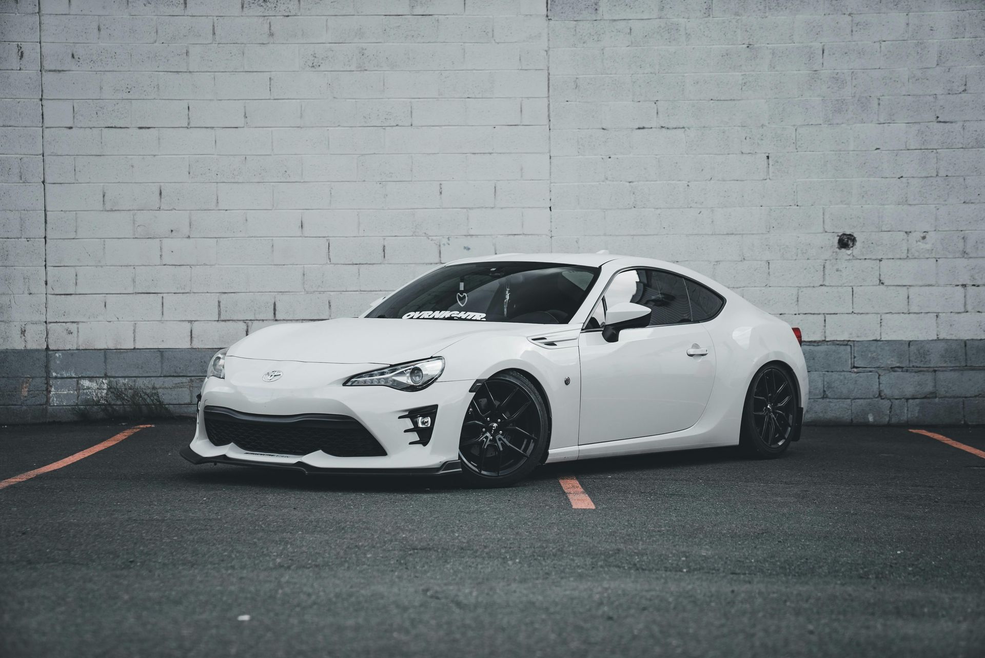 White sports car parked in front of a brick wall; black wheels and front lip.