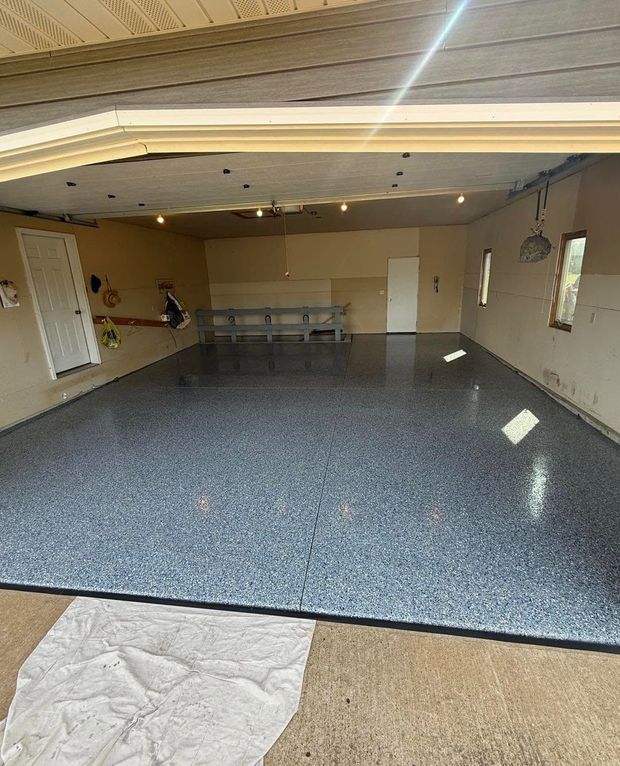 Empty garage with speckled epoxy floor, beige walls, ceiling lights, and a workbench at the back.