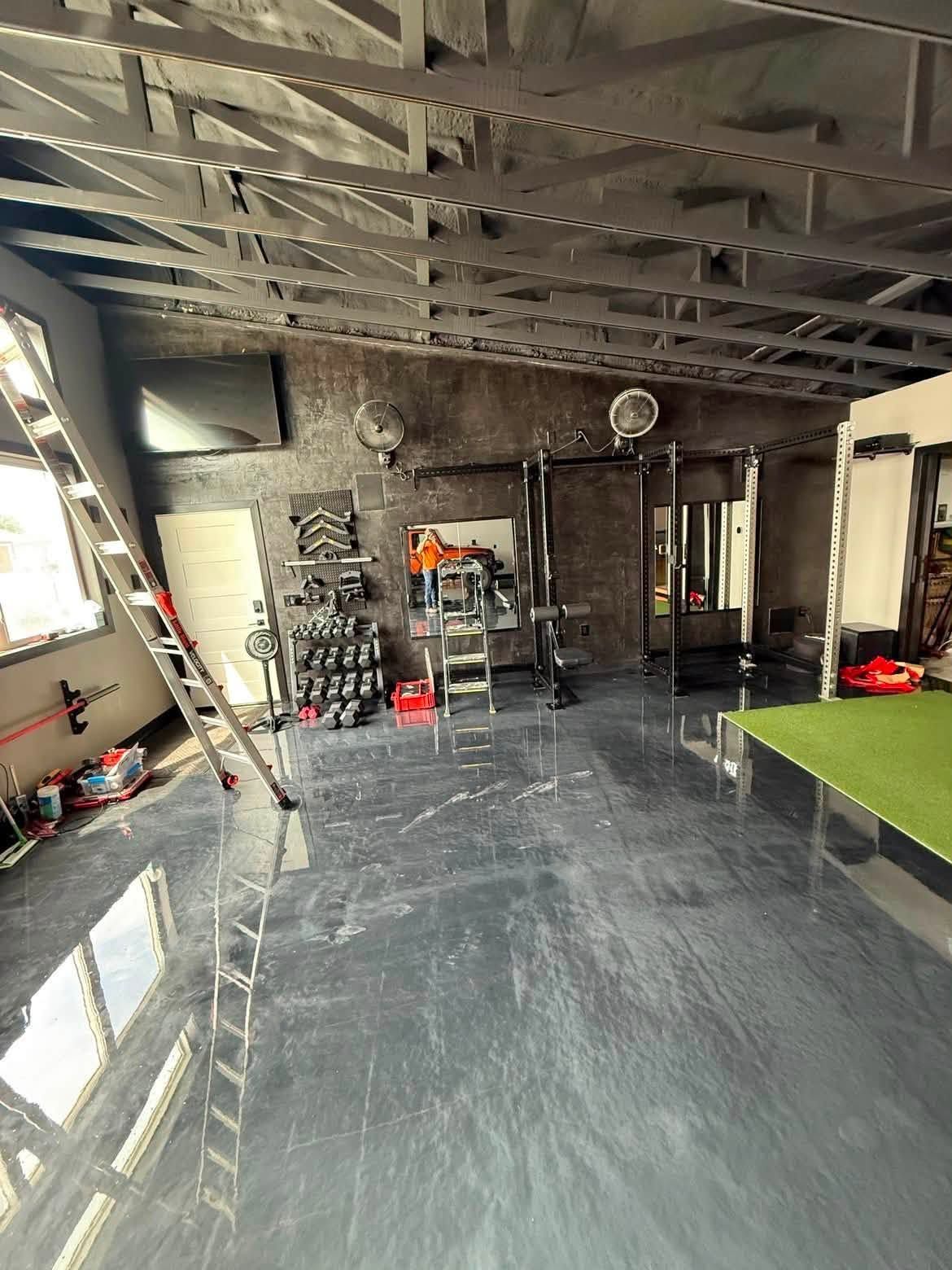 Indoor garage with glossy gray floor, exposed rafters, ladder, tools, and a green mat by the right wall