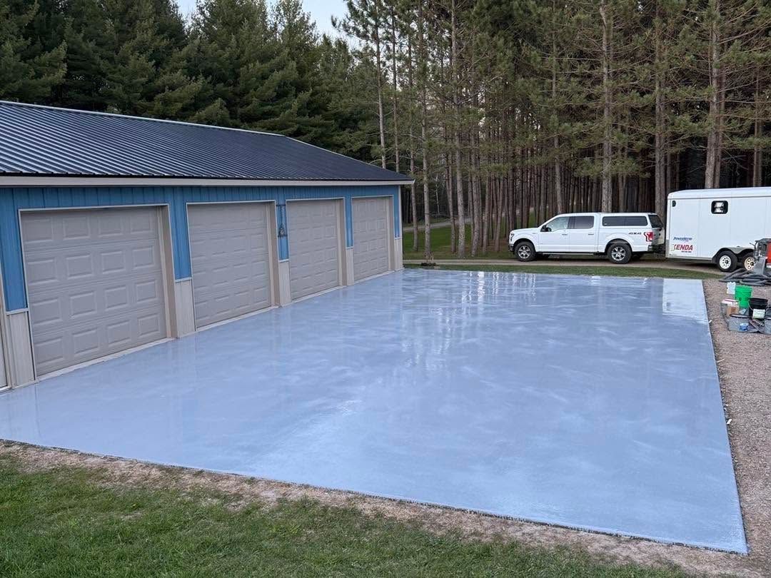 Blue epoxy driveway beside a garage, with a white pickup and trailer parked near a wooded area.