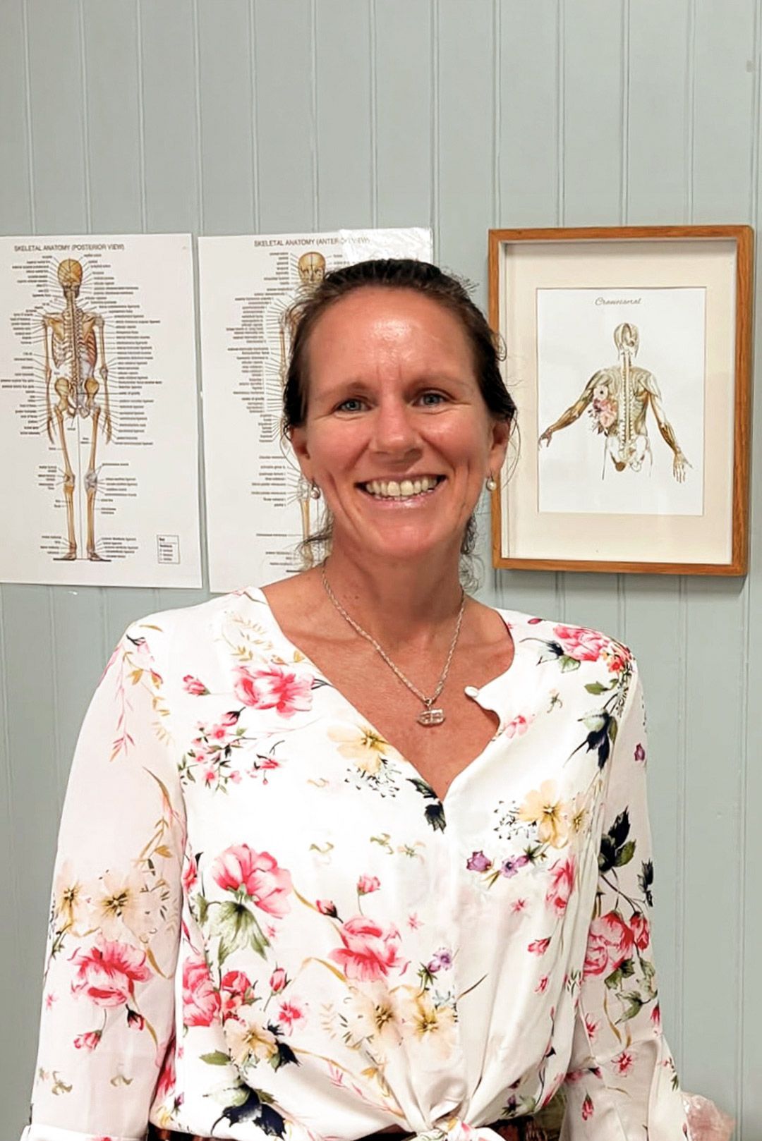 Christine Hollis - Biodynamic Craniosacral Therapist Photo of lady wearing white floral sirt with pink flowers and medial diagram of skeleton hanging on wall behind.