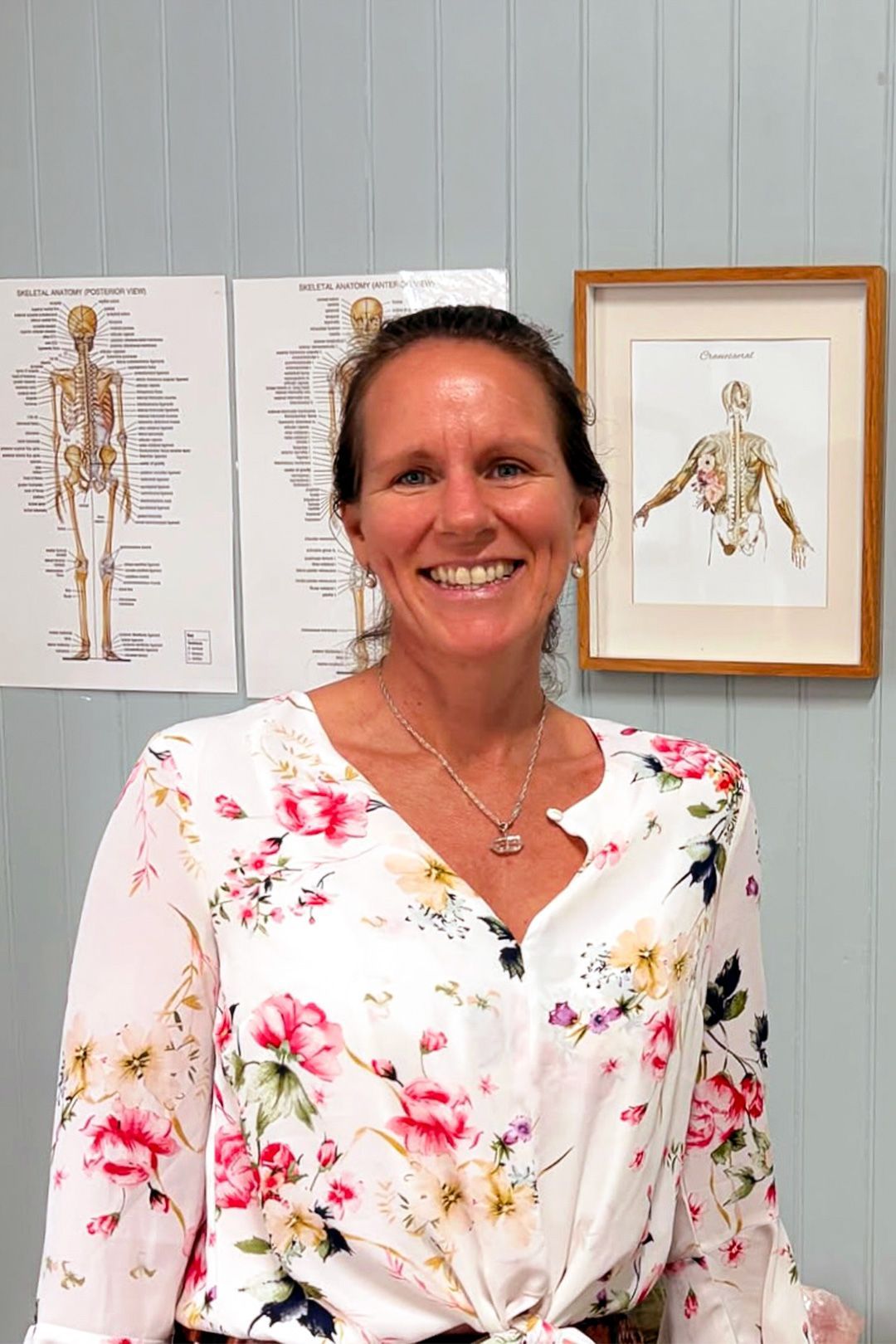 Photo of lady wearing white floral sirt with pink flowers and medial diagram of skeleton hanging on wall behind.