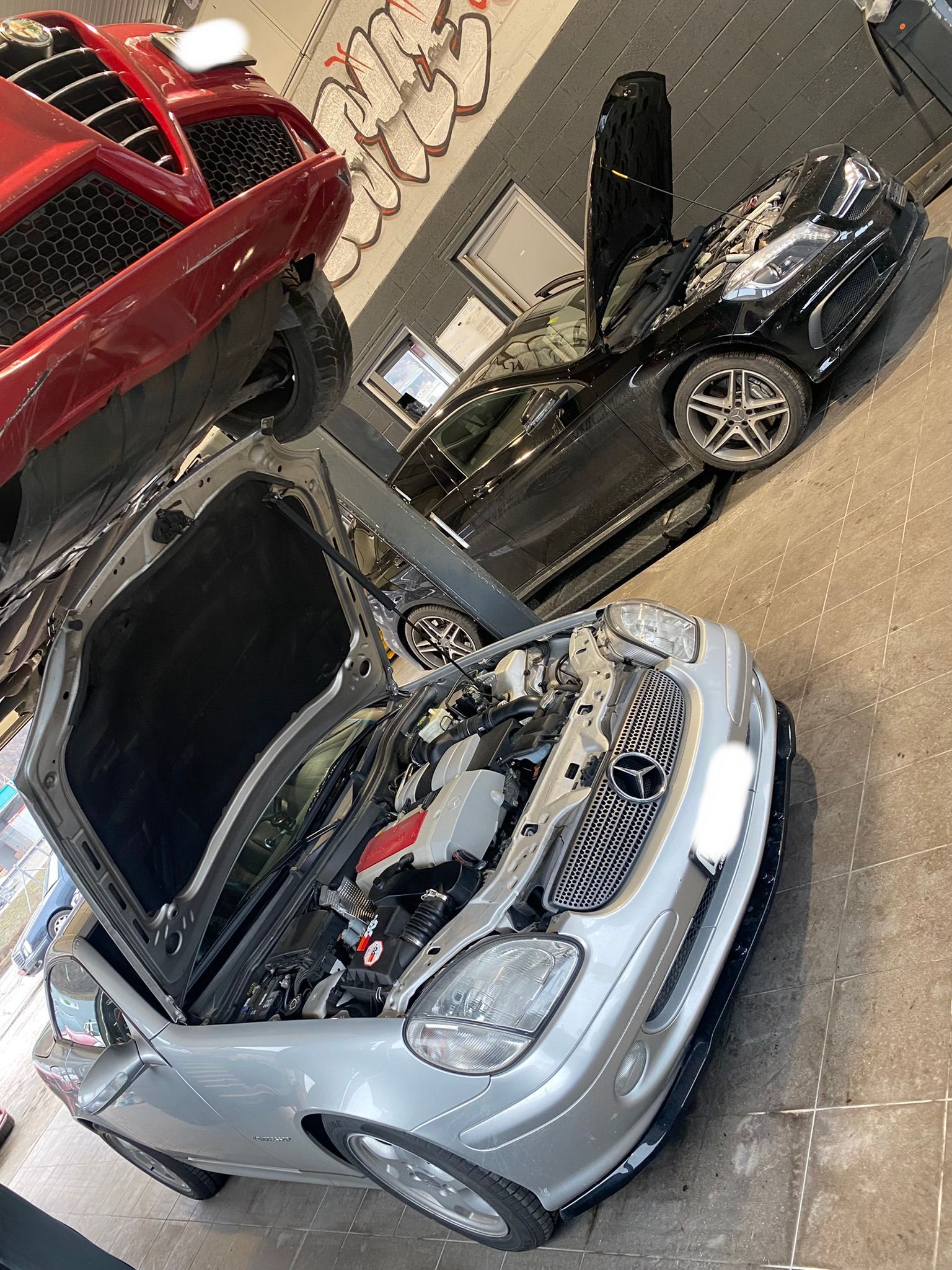 Une voiture argentée avec son capot ouvert dans un garage à côté d'une voiture rouge.
